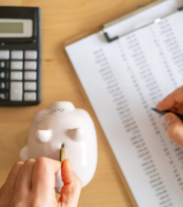 Accountant inserting a coin into piggy bank and calculating expenses with calculator and spreadsheet on a wooden desk, managing home budget and savings