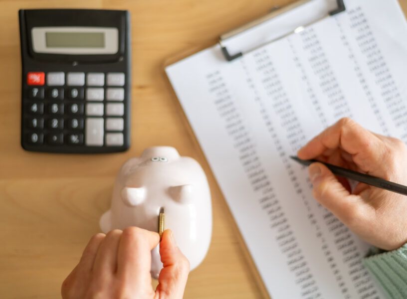 Accountant inserting a coin into piggy bank and calculating expenses with calculator and spreadsheet on a wooden desk, managing home budget and savings Accountant inserting a coin into piggy bank and calculating expenses with calculator and spreadsheet on a wooden desk, managing home budget and savings